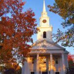 Historic white church with steeple surrounded by vibrant autumn trees under a clear blue sky.