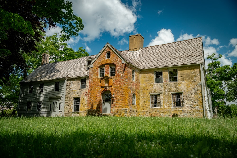 An external view of Spencer-Peirce-Little farm. It is a partly cloudy day and green grass is visible with a shadow being cast over part of it by a large tree.