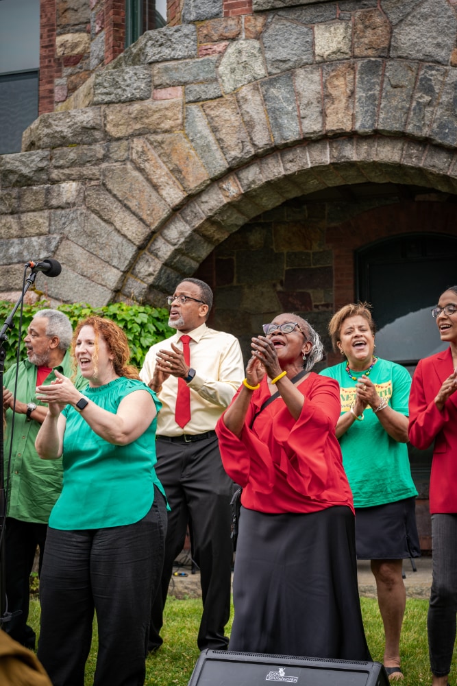A group of people, standing in front of a stone arch at Eustis perform as part of a Juneteenth community celebration.