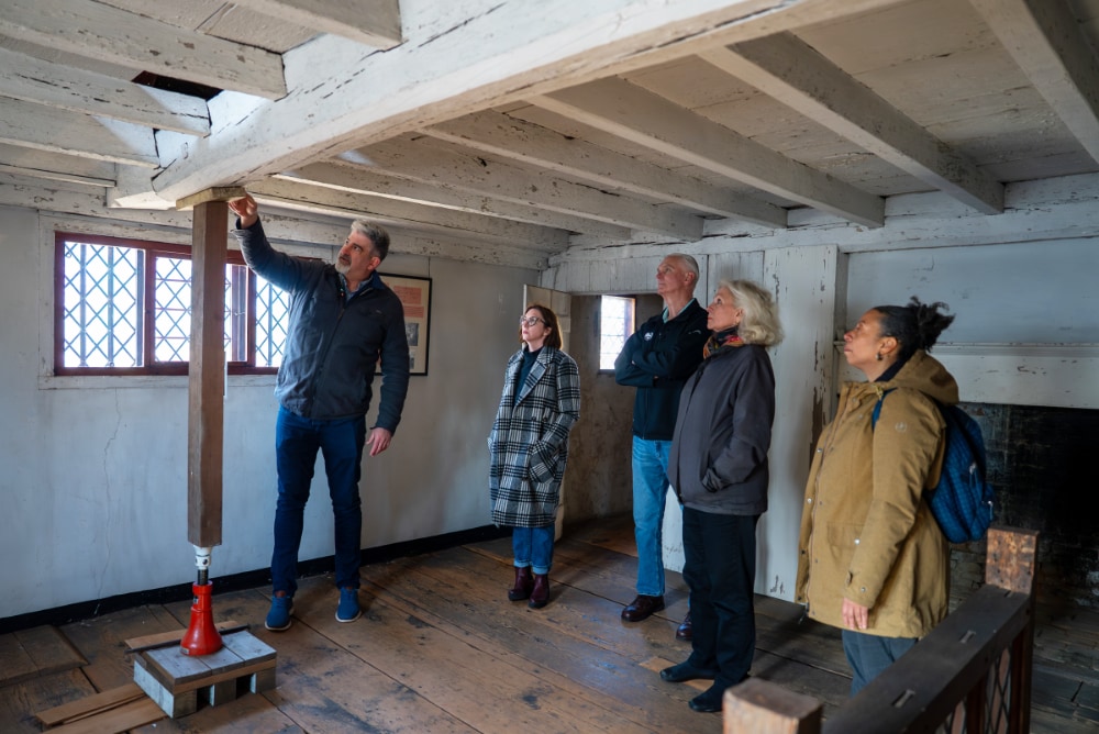 A tour guide at Browne House points out a support in a room going from the floor to a beam in the ceiling.
