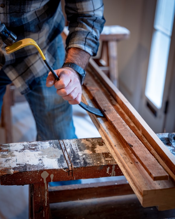 A woodworker uses a pry bar and hammer on a large piece of wood at Pierce House.