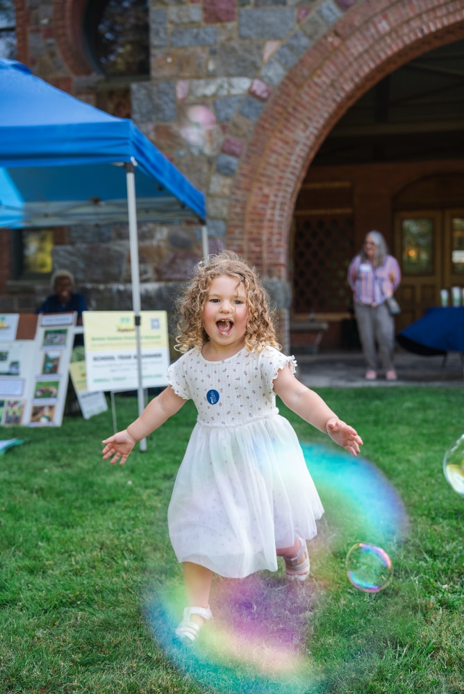 A young girl, laughing, running in the grass amongst bubbles. She has curly blonde hair and is wearing a white dress and sandals.