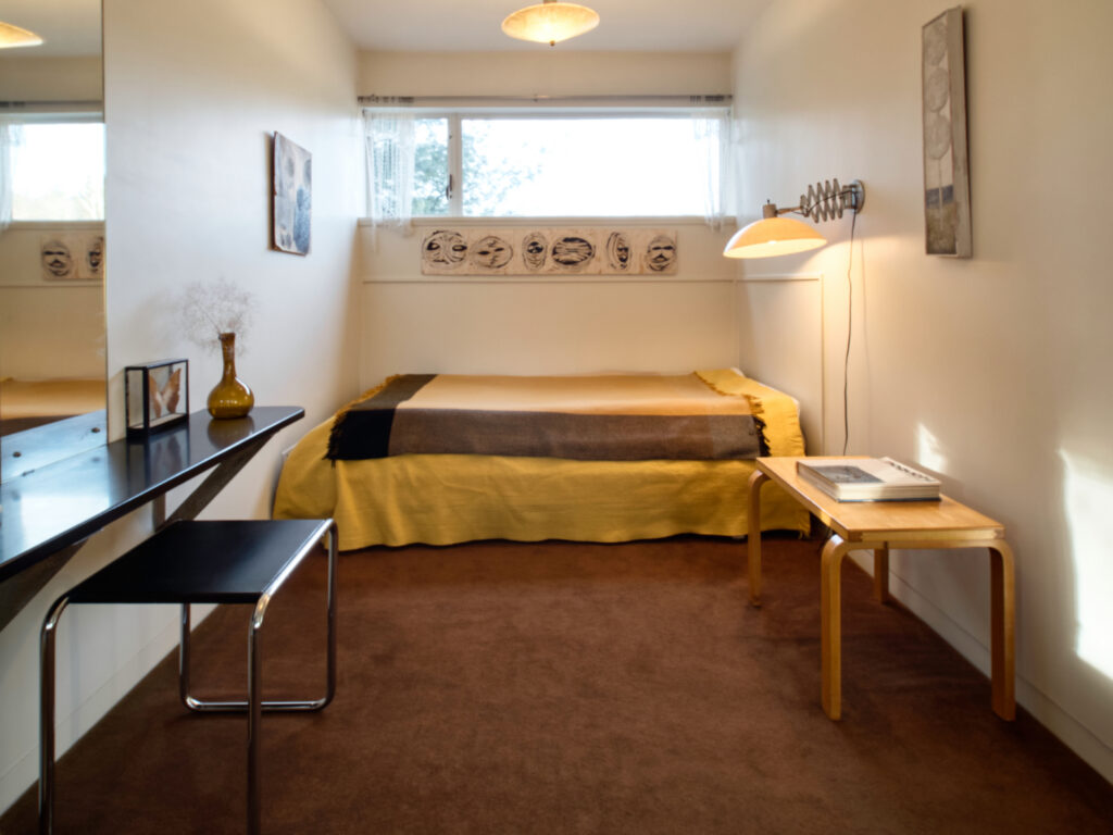 A sleeping nook in Gropius House showing a bed, lamp, bench with counter and an end table.