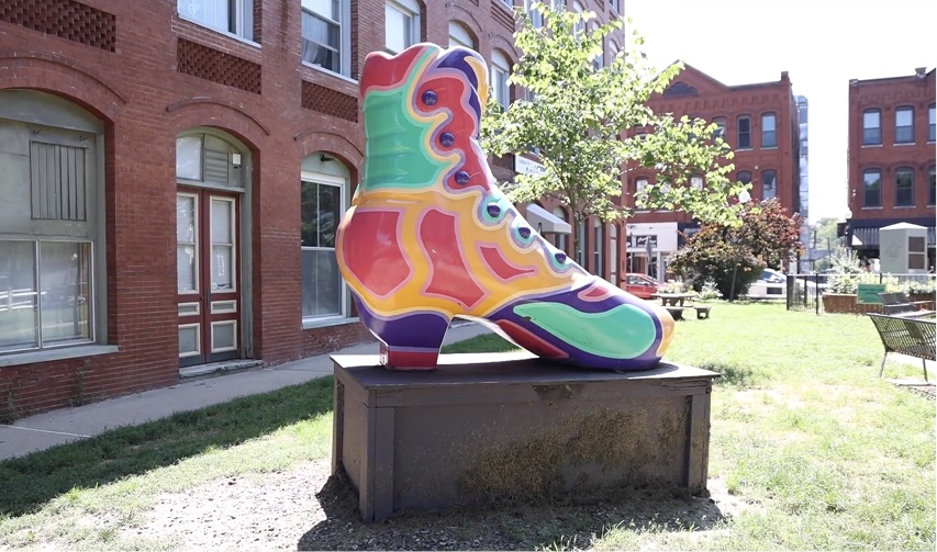 A photo of a large, colorful shoe sculpture on top of a pedestal. The shoe is in a park surrounded by brick buildings.