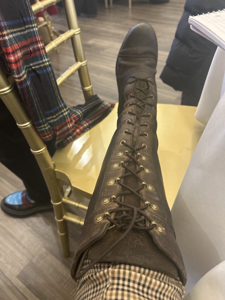 Brown leather lace-up boot with brass eyelets and embossed logo, worn by a seated person in a social indoor setting with a gold chair and plaid scarf in the background.