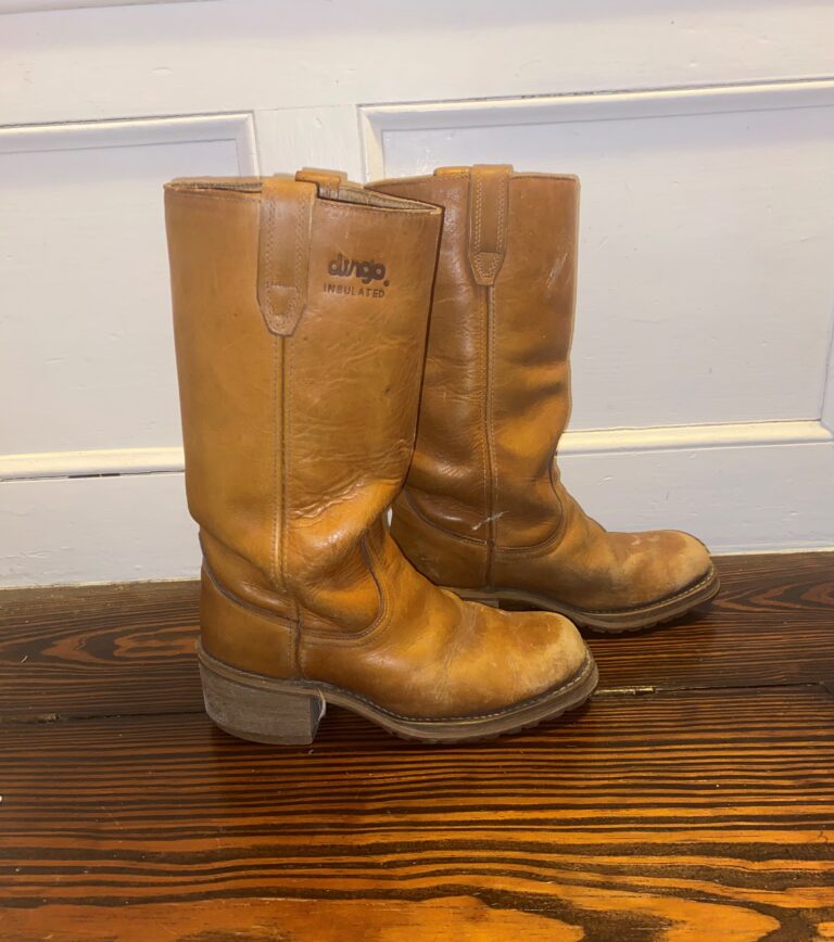 Worn brown leather mid‑calf boots with visible creases and scuffs, resting on a wooden floor against a white paneled wall.