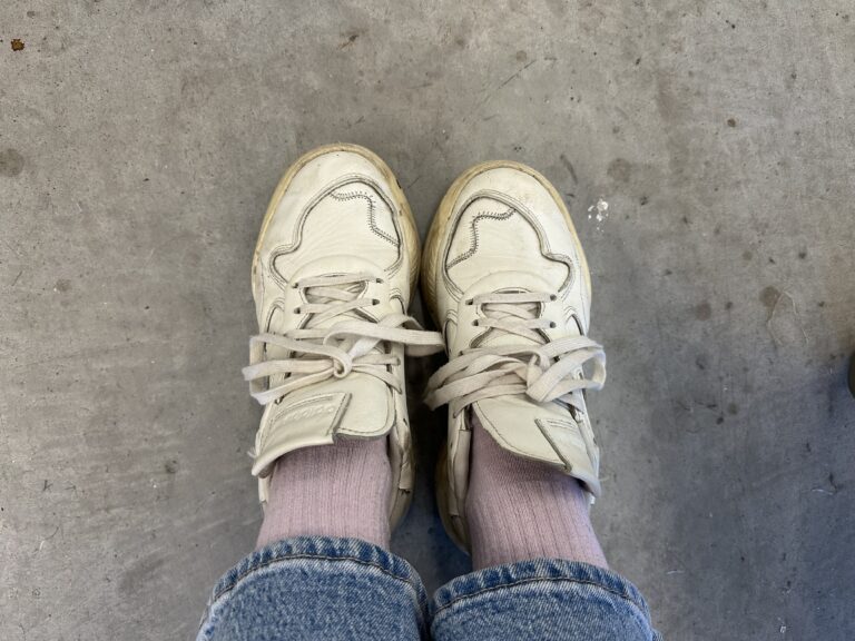 White sneakers with light wear, worn with pink socks and cuffed blue jeans while standing on a concrete floor.