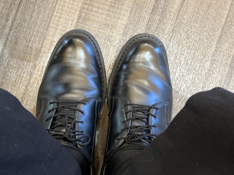 Black leather dress shoes with visible toe creases, worn with black socks and dark pants on a wood-patterned floor.