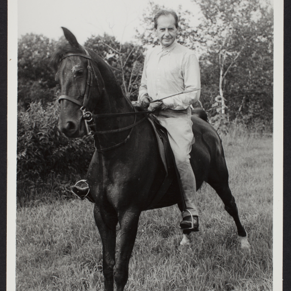 A tall, lanky white man sits on the back of a standing horse. The man is wearing riding boots that fit into the stirrups.