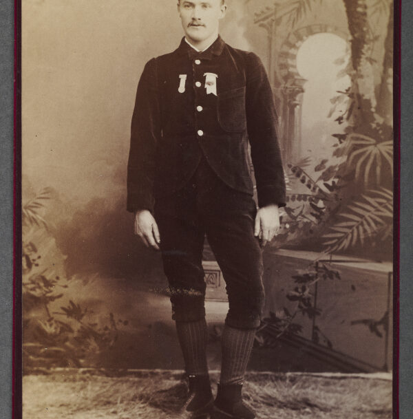 Professional sepia tone photographic portrait of a White man in a dark velvet cap, matching, dark, long sleeve button up jacket and short, knee length pants with tall, thick knitted socks, and roller skates. He wears award ribbons on his jacket front and stands against a backdrop with an imagined orientalist architecture and plant landscape.