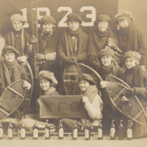 A sepia tone group portrait of thirteen young, White college women dressed in winter hats and scarves holding either snowshoes or skis. Behind them is a large fabric banner reading 1923 and two women in front hold a banner with the Greek letters chi, gamma, theta.