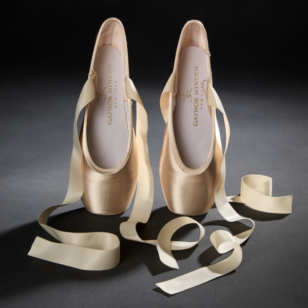 A pair of light creamy pink satin pointe shoes resting vertically on their toes with the insoles facing the viewer and wide satin ribbons streaming down around the shoes.
