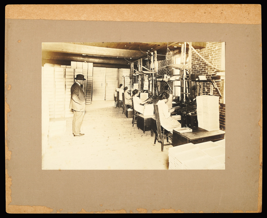 Sepia tone photograph of a factory building interior with hundreds of stacked boxes in the background and a middle aged, White man standing in a suit and a hat overlooks and group of four White women with dark hair all wearing dresses and sitting down at machinery along a brick wall with windows.