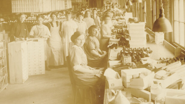 A sepia tone photograph of a factory interior with windows on the right and a row of seating women looking at the camera with shoes in front of them and more workers standing in the back looking at the camera.