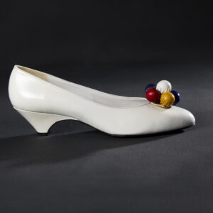 Side profile of a white leather woman’s shoe with a curved, low heel adorned with multicolor leather covered spheres.