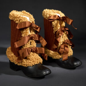 Two boots with shiny black rubber bottoms and brown knitted yarn shanks adorned with curly looped yarn and three brown, satin ribbon closures siting side by side.