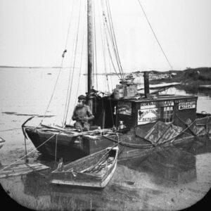 A black and white photograph of an older White man with a beard on a small wooden sailboat off the coast with a small dog sitting below advertising signs reading “W. O. Cottle Boots and shoes repaired,” “Scow. was built by W. O. Cottle 1896, Yankee Notion, Hom eon the Ocean, Boots and Shoes Repaired.”