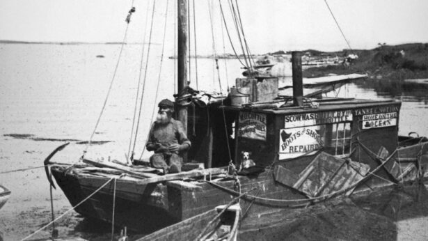 A black and white photograph of an older White man with a beard on a small wooden sailboat off the coast with a small dog sitting below advertising signs reading “W. O. Cottle Boots and shoes repaired,” “Scow. was built by W. O. Cottle 1896, Yankee Notion, Hom eon the Ocean, Boots and Shoes Repaired.”