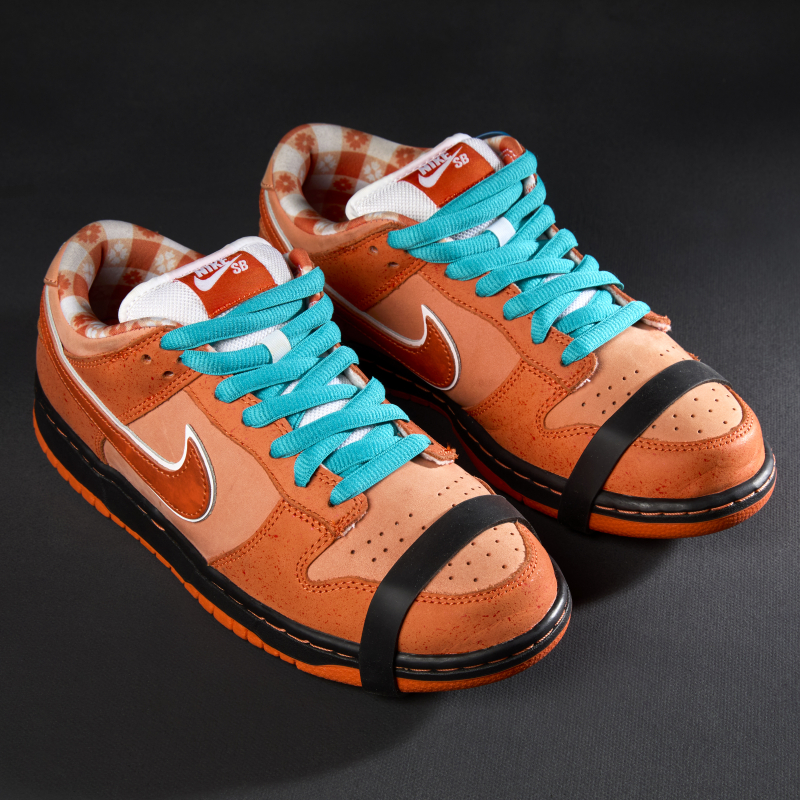A pair of multi-tone orange suede and leather sneakers with large Nike swoop on side, orange and white picnic table print interior, and black and orange rubber soles, bright blue shoelaces, and a rubber lobster claw style band on around the toe.