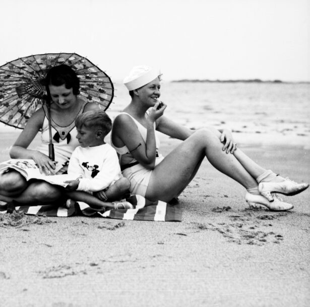 Two White women site on either side of a young boy on a beach on a blanket with thick stripped pattern. One holds a parasol and looks down at a magazine the other is putting on lipstick, turned to the side and facing away from the camera and wears a sailor hat. All are wearing shoes.