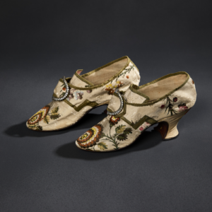 A pair of light-colored heeled silk shoes with elaborate floral multicolored embroidery and rhinestoned buckles.