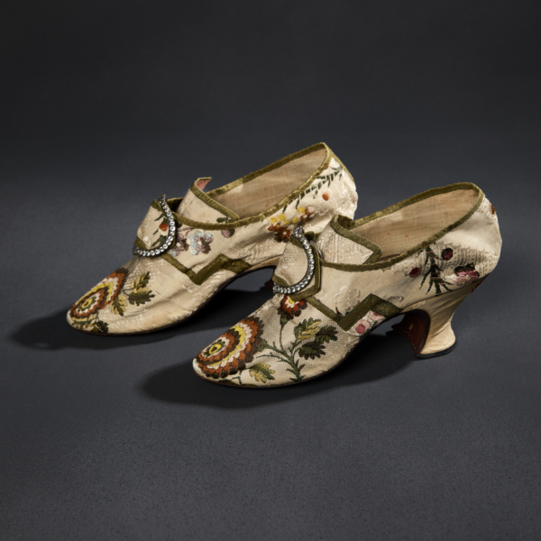 A pair of light-colored heeled silk shoes with elaborate floral multicolored embroidery and rhinestoned buckles.