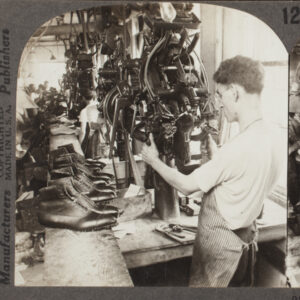 Two of the same images side by side and mount on paper reading “Keystone View Company, Copyrighted, Manufacturers, Made in the U.S.A., Publishers” and “22189 Lasting Machine Shaping Shoes in a Massachusetts Shoe Factory.” Image is a black and white photograph of a thin, White man using a large machine in a factory with his back to the viewer and a line of shoes on a work bench to the left.