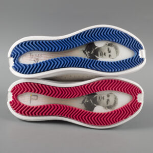 Two white sneakers stacked vertically on top of one another with the bottoms facing the viewer. Top shoes sole is outlined in red and the bottom shoe’s sole is outlined in blue. The center of each sole has a picture of Jan E. Matzeliger, an Afro-Surinamese inventor along with small writing.