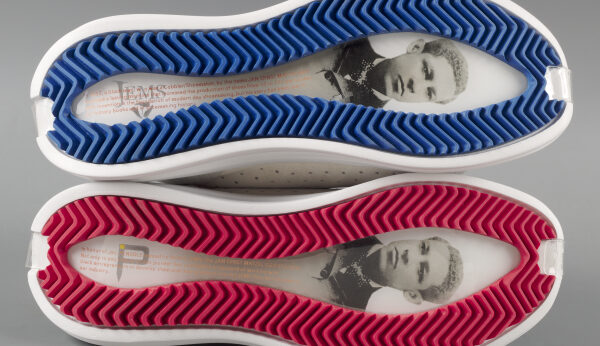 Two white sneakers stacked vertically on top of one another with the bottoms facing the viewer. Top shoes sole is outlined in red and the bottom shoe’s sole is outlined in blue. The center of each sole has a picture of Jan E. Matzeliger, an Afro-Surinamese inventor along with small writing.