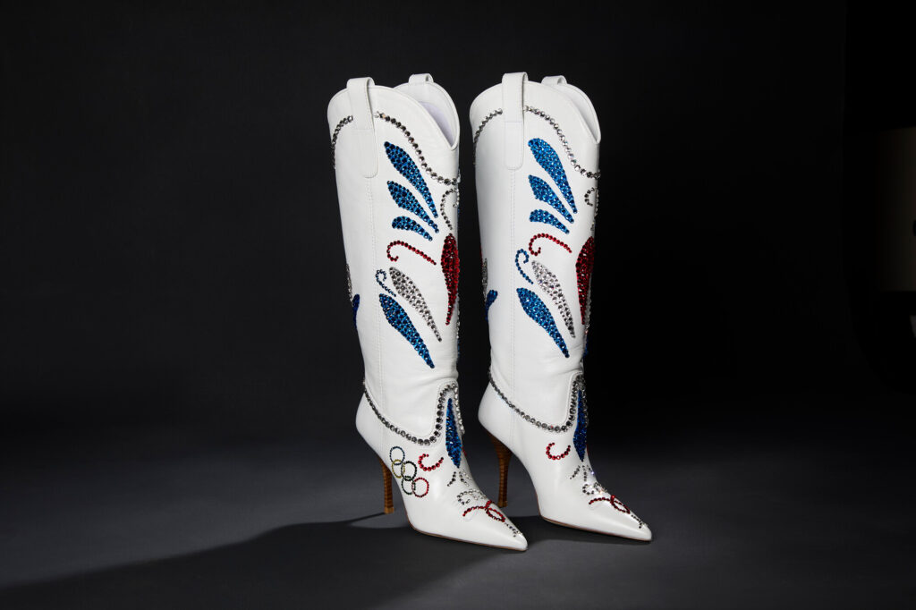 A pair of white leather knee high cowboy style boots with a pointed toe and stiletto heel adorned with silver, red, and blue rhinestones in traditional floral pattern and Olympic ring logo on the side of the foot.