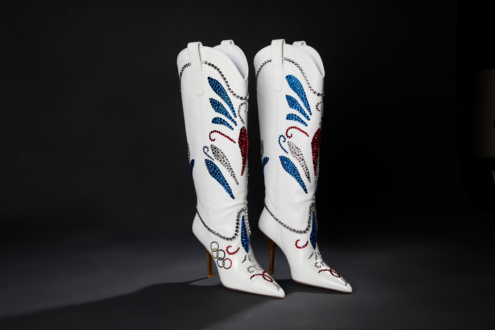 A pair of white leather knee high cowboy style boots with a pointed toe and stiletto heel adorned with silver, red, and blue rhinestones in traditional floral pattern and Olympic ring logo on the side of the foot.