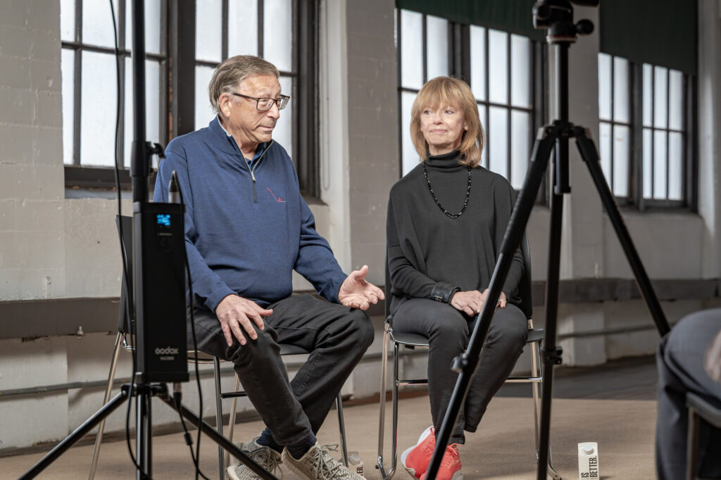 An older White man with glasses and wearing a blue, long sleeve zip up, black pants, and grey sneakers sits with an older White woman with shoulder length strawberry blonde hair and bangs wearing a black turtleneck shirt, black beaded necklace, black pants, and red sneakers. They are being interviewed and filmed in a former factory building room with white walls and large windows.