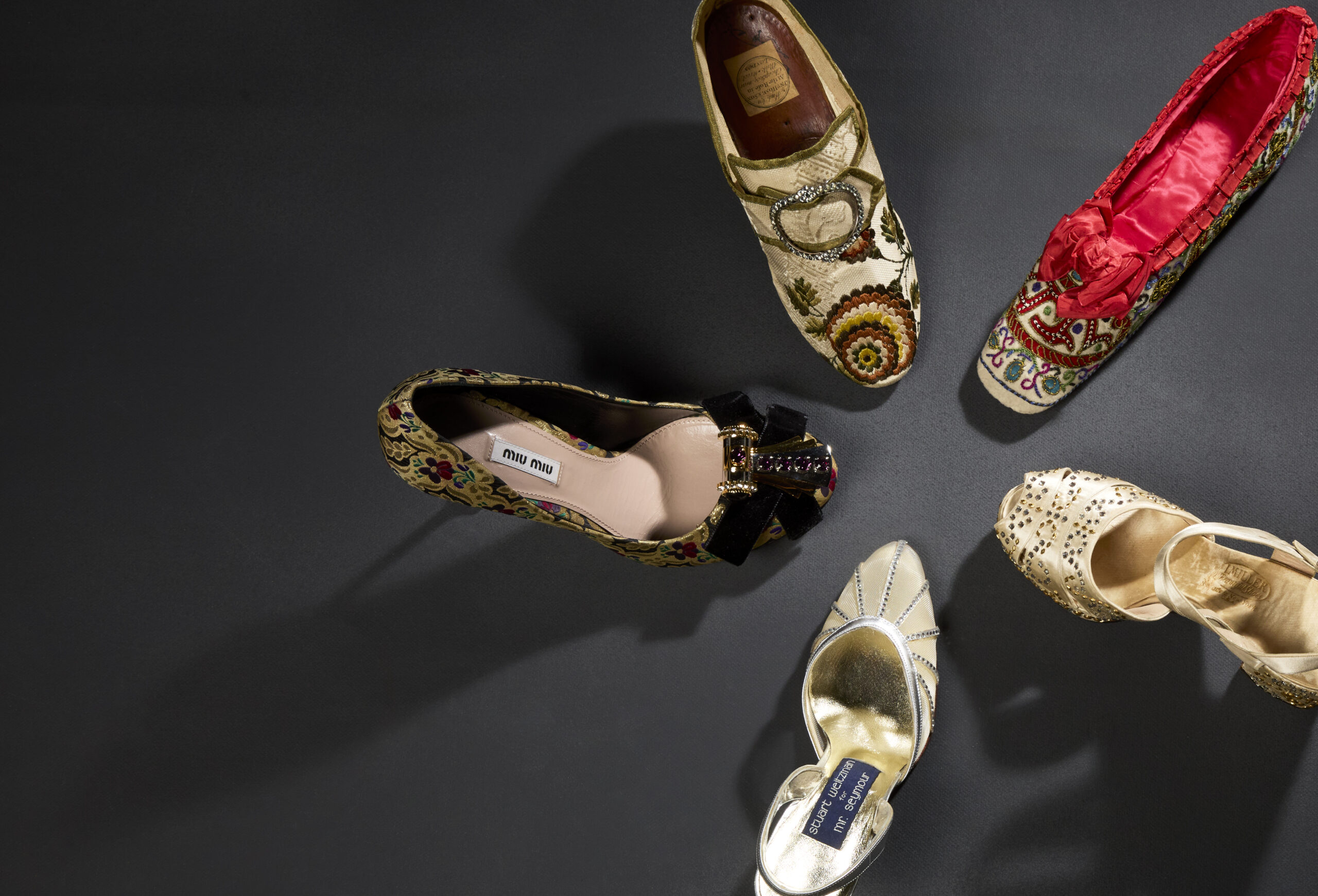 A circle of five women's shoes. Clockwise: eighteenth century, English, silk brocade slipper with buckle closure, 1860s embroidered slipper with red silk lining, 1940s I. Miller platform sandal with metallic embellishment, 1980s Stuart Weitzman sling-back heel, 2010s Miu Miu brocade pump with velvet bow and shoe clip.