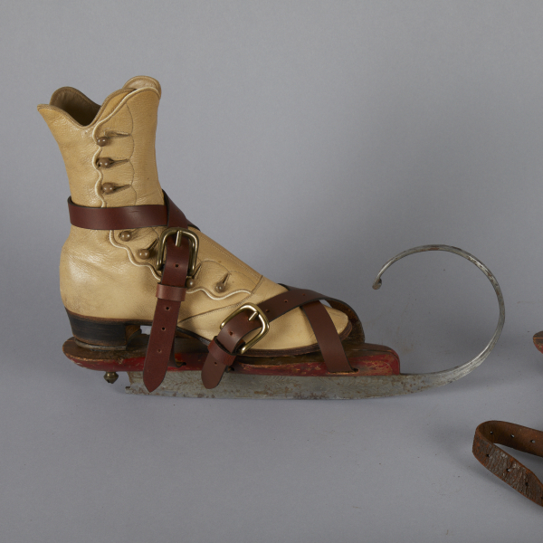 Two antique wooden ice skates with large, silver metal curves coming off the blades on the bottom. Each has leather straps, and one has the straps wrapped around a tall, tan leather, button top boot to secure it to the skate.
