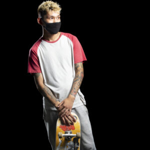 Color photograph of a young Asian man, with blonde styled hair, and multi-colored tattoos on both arms wearing acid wash, wide-leg jeans, a white ringer-tee with red short sleeves and collar, white sneakers with black sneakers, and a black fabric face mask covering his nose and mouth, standing against a black background. He leans his weight on his back, right foot and standing with his hands crossed, holding to the top of his skateboard with the wheels pointed toward the camera.