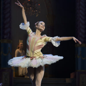 Photograph of a White, woman with blonde hair pulled back in a bun, dancing ballet en pointe, wearing a yellow and pink stripped bodice and stiff white tutu adorned with flowers and ribbons.