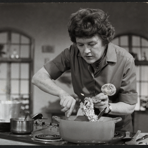 A black and white photograph of a woman with curled hair wearing a collared shirt standing over a stovetop with several pots and pans. Behind her are two large windows.