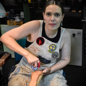 A white woman with dark hair stares at the camera. She is seated in a workshop, wearing a well-worn apron covered in iron-on patches and using a tool on the bottom of a shoe held in her other hand.