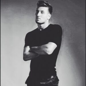 A young white man wearing dark clothing looks at the camera with his arms crossed. His head and body are turned slightly so that part of his face and body are in shadow.