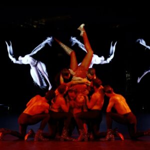 A group of shirtless male dancers carries a female dancer who dances upside down wearing a red bodysuit, a cantilever heel on her left leg, and a spike prosthetic on her right leg. In the background, digital graphics morph between human and imagined shapes.