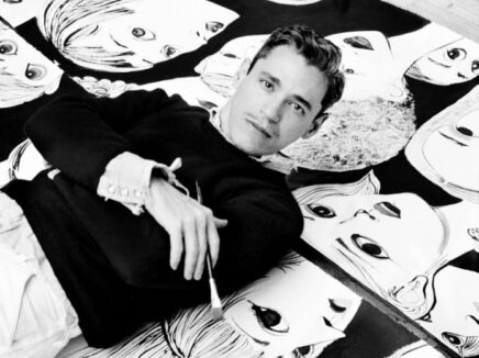 Black and white photo of a man looking into the camera while holding a paintbrush and lying on a background patterned with stylized graphics of faces.