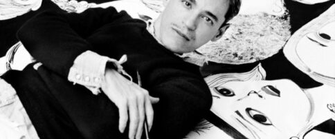Black and white photo of a man looking into the camera while holding a paintbrush and lying on a background patterned with stylized graphics of faces.