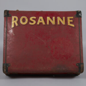 A red painted metal case, well-worn and with some scratches, scuffs, and rust. “Rosanne” painted in thick, bold, capital letters in yellow and white, with a dark red shadow.