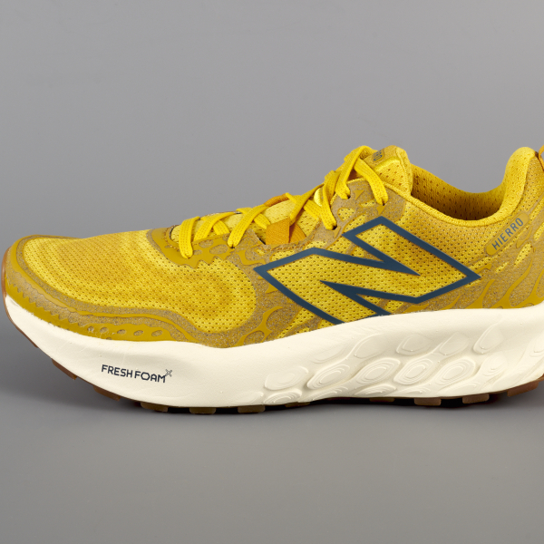 A single sneaker in side profile with a mustard yellow, highly textured top and large outline of an “N” in dark blue, creamy white mid-soles, and translucent orange-brown bottom soles.