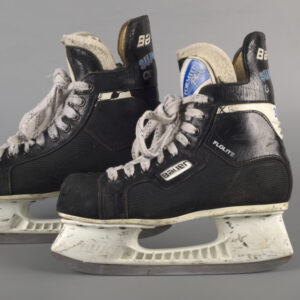 A pair of well-worn modern hockey skates in side profile and leaning against one another. Black leather tops with white laces, “Bauer” on the side, and white, scuffed, plastic blade guards above thick silver colored metal blades.