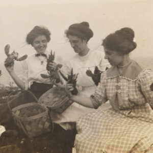 A group of four women, in long dresses with their hair up but windswept, hold lobsters in each hand. They are gathered around a basket with the ocean in the background, smiling and looking at the lobsters.