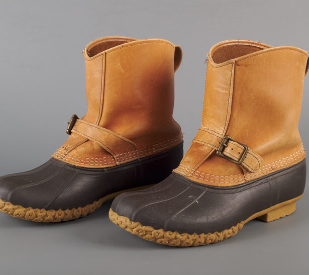A pair of iconic duck boots, with a tan textured sole, a black rubber lower, and a leather upper. There are three lines of visible white stitching connecting the rubber to the leather, and a buckled strap across the leather on each boot.