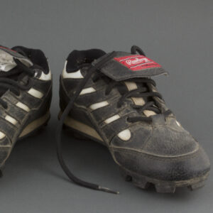 A pair of small, worn, black baseball cleats with white accents. The laces are black and the tongue is black with a red label reading Rawlings.