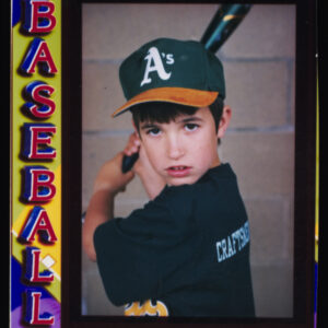 A mocked-up baseball card with a photograph of a young boy posed with a bat in his Little League uniform. Alongside the photograph reads, "Baseball 98 Supercard" on a colorful background.
