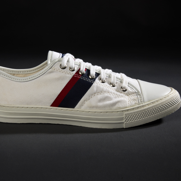 A side profile photograph of a single white canvas sneaker, with thick diagonal band of dark blue and red on the side, white rubber sole, and white cotton laces.